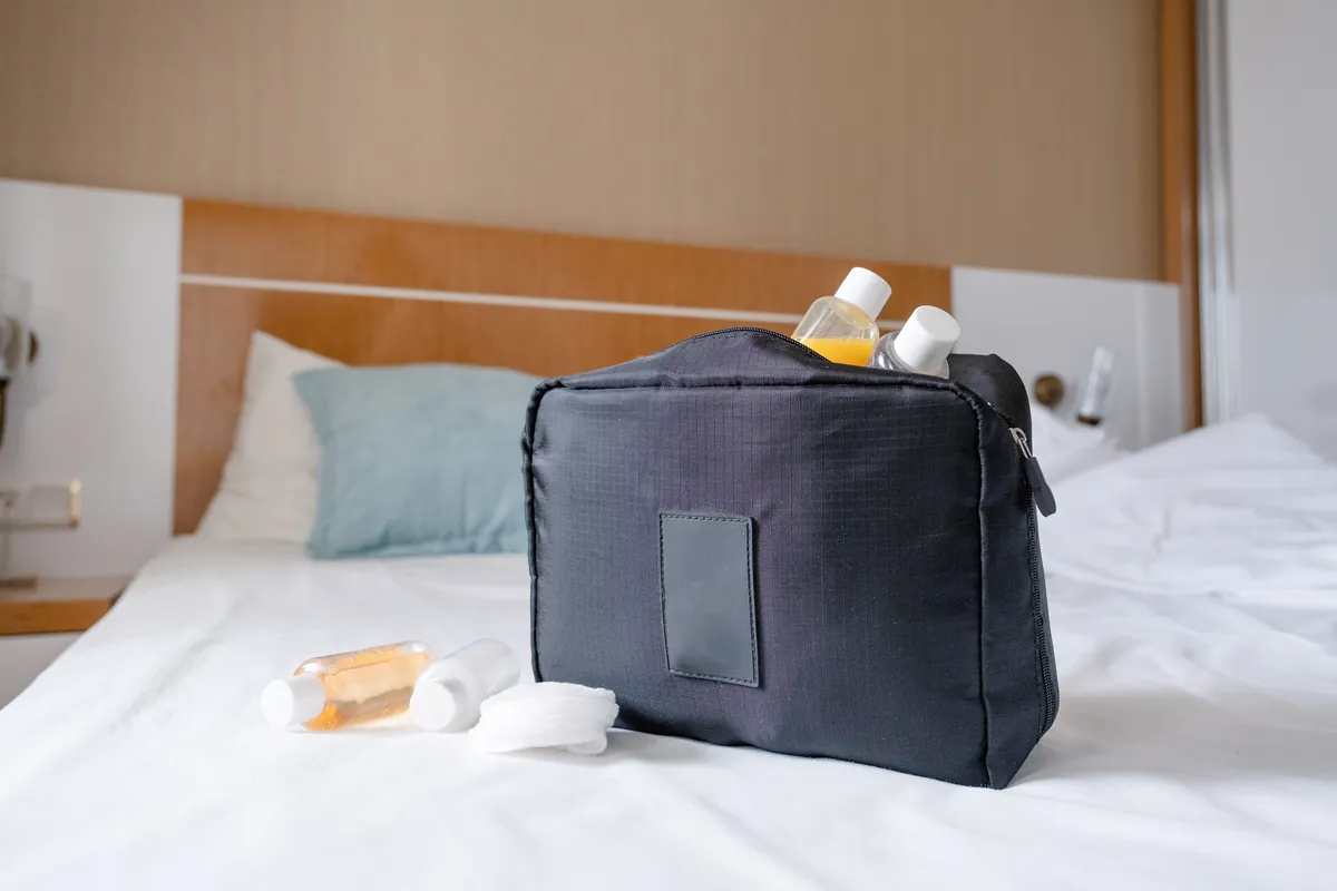 Black zippered toiletry bag standing on a white bed, with small travel bottles and cotton pads beside it