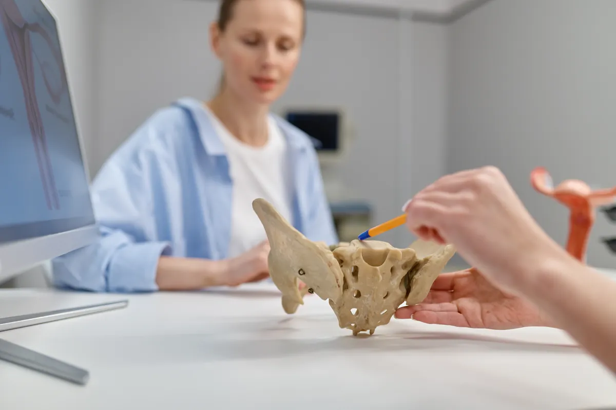 Clinician pointing with a pen at a model of the bony pelvis on a white desk, while a woman in a light blue shirt watches in the background next to a computer screen