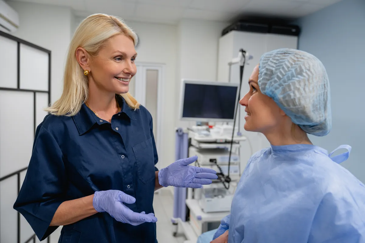 Smiling woman in navy scrubs and purple gloves talking with a woman in a blue disposable gown and surgical cap in a clinical room