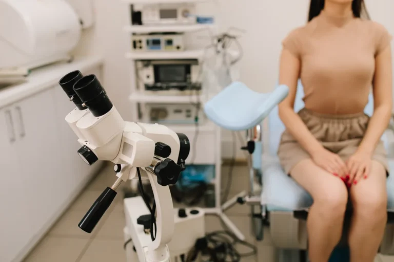 Colposcope in a gynecology exam room, with a clothed patient sitting on the exam chair in the background
