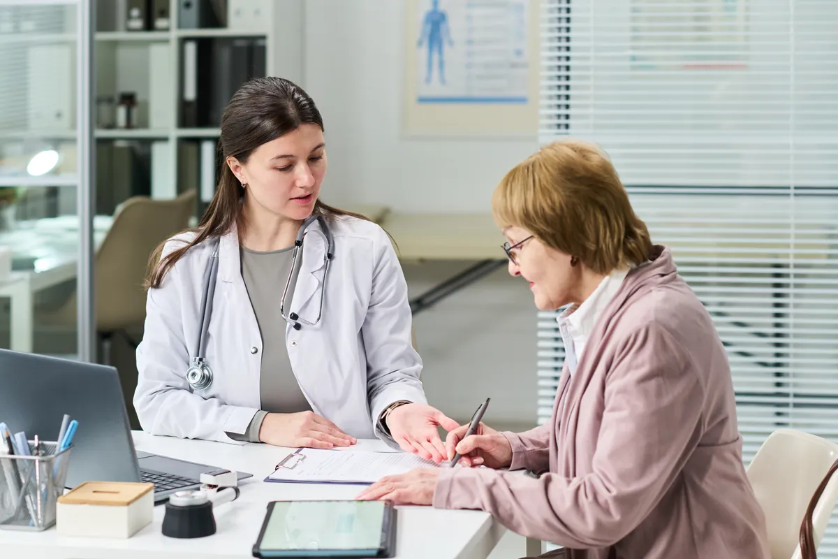 Young doctor in a white coat with a stethoscope explaining a form to an older woman with glasses and a pink jacket who holds a pen at a desk
