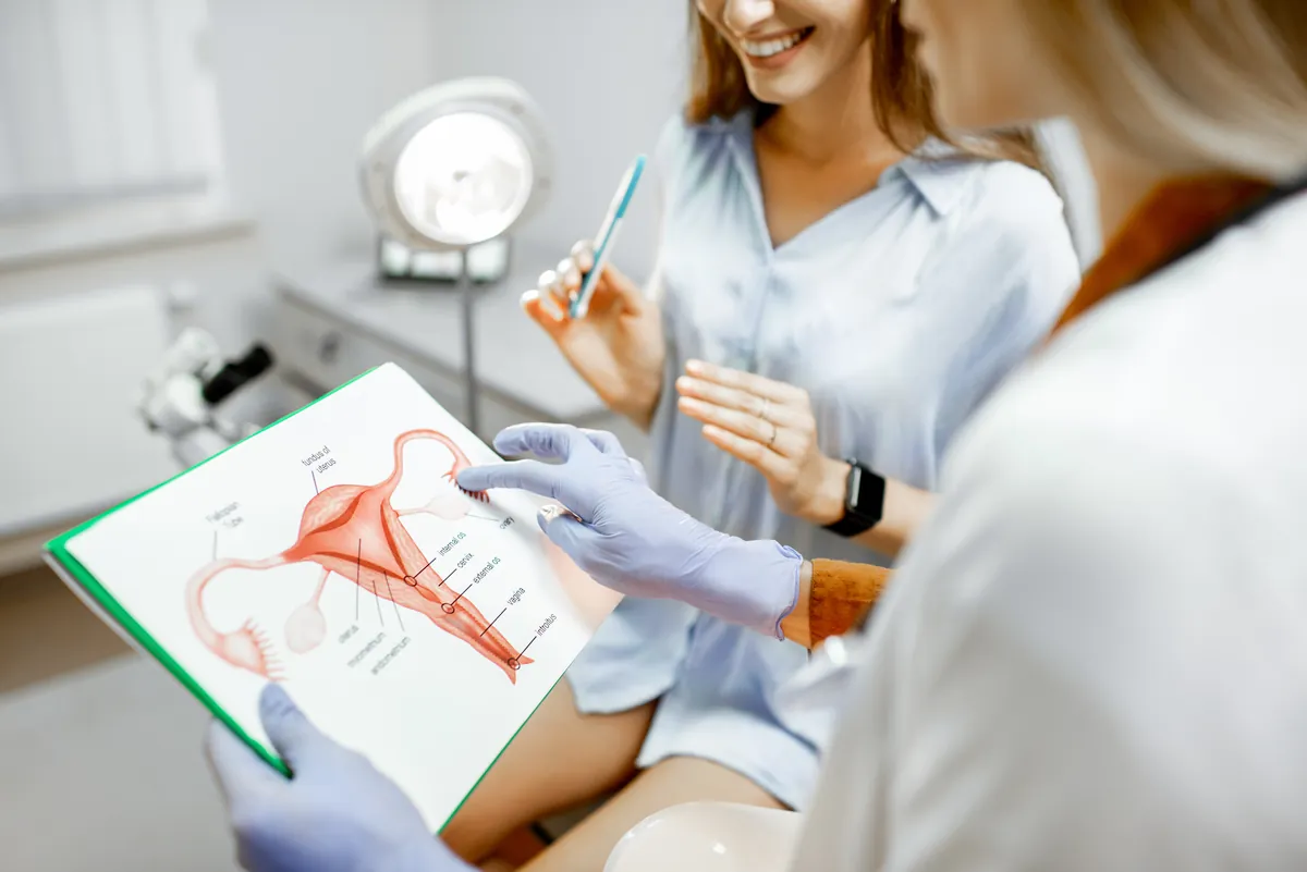Gloved doctor pointing to the end of the fallopian tube next to the ovary on a labeled drawing of the female reproductive organs while a smiling patient listens