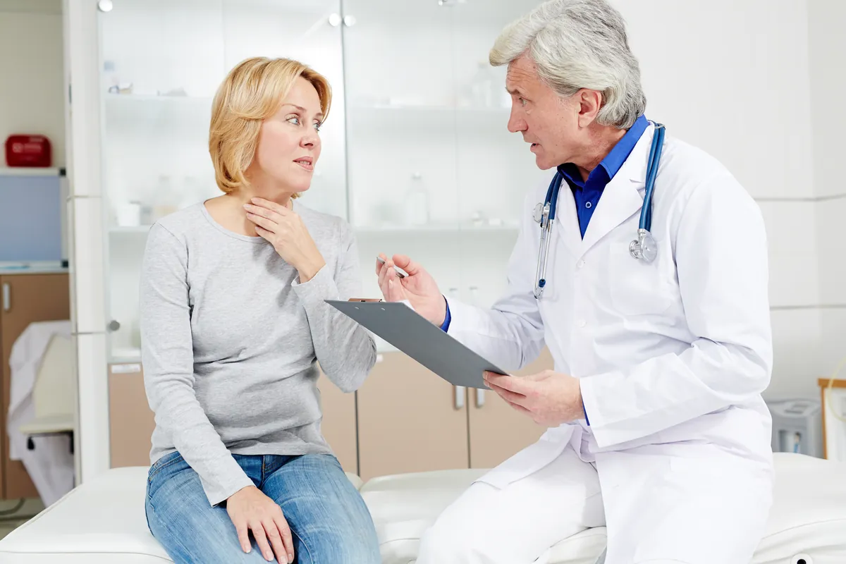 Grey-haired doctor with a stethoscope and a clipboard talking with a blonde woman in a grey top who sits on an exam table with her hand at her neck