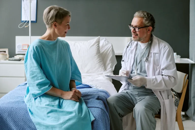 Doctor with glasses, a white coat and gloves holding a clipboard and talking with a smiling woman in a turquoise gown sitting on a hospital bed