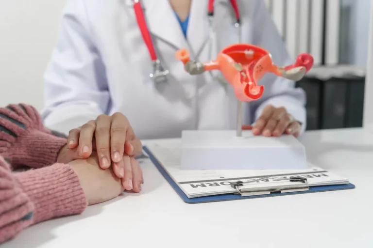 Doctor in a white coat with a model of the uterus on the desk, facing a patient in a pink sweater with her hands clasped