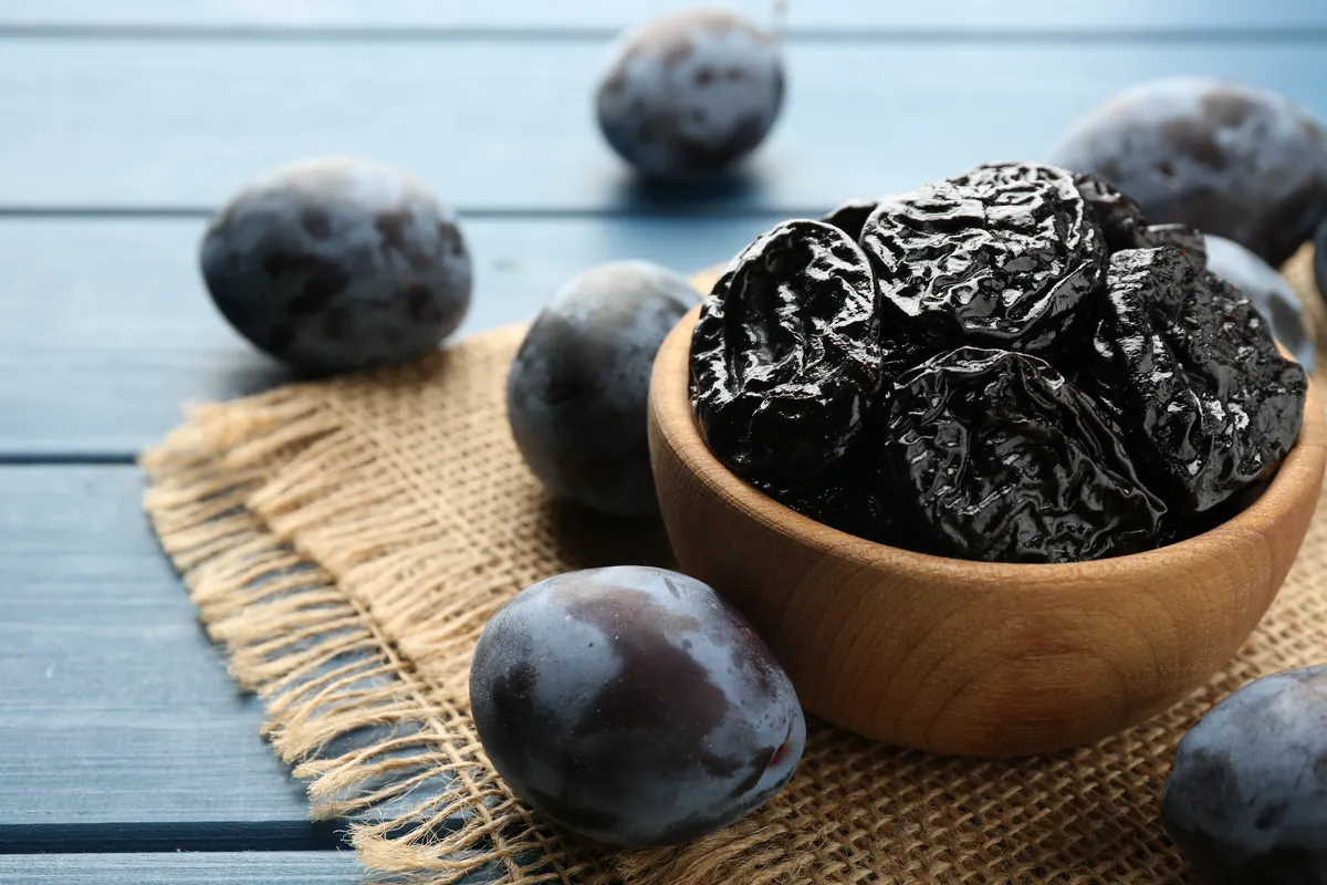 Wooden bowl of dried prunes on a burlap cloth, surrounded by fresh plums on a blue wooden table