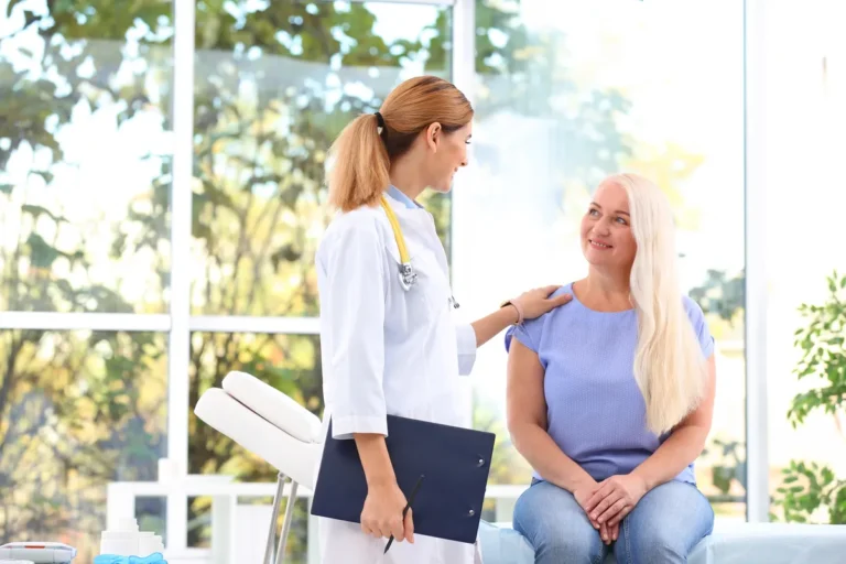 Female doctor in a white coat holding a clipboard rests a hand on the shoulder of a smiling middle-aged woman with long blonde hair sitting on an exam couch by large windows