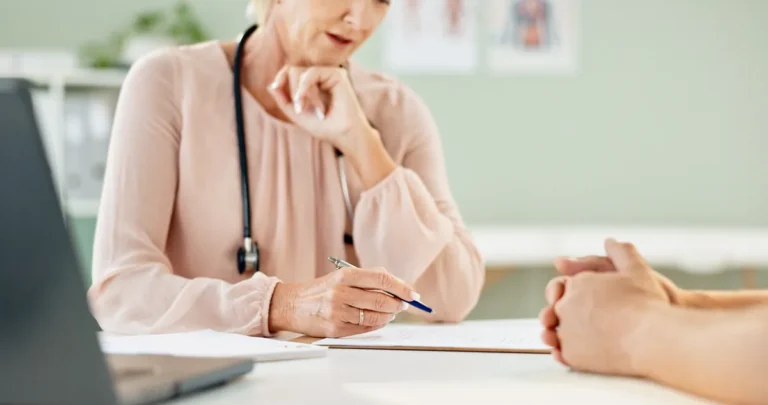 Female doctor with a stethoscope and a pale pink blouse taking notes at a desk while listening to a patient whose clasped hands rest on the table