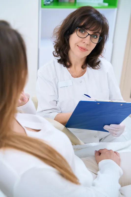 Doctor with glasses and a white uniform holding a blue clipboard and smiling while she listens to a patient seen from behind