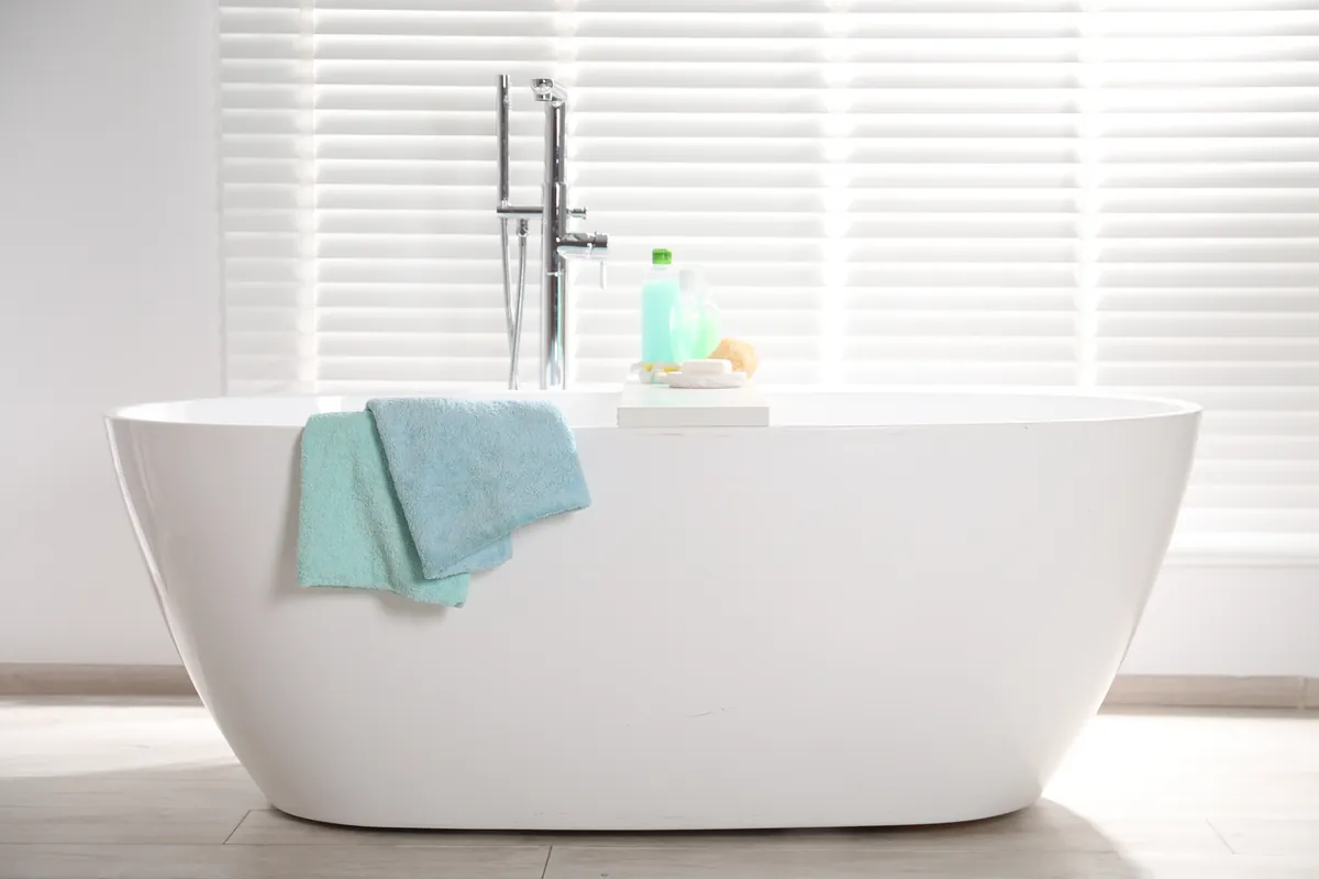 Empty white freestanding bathtub with two teal towels draped over the side, in front of a window with white blinds