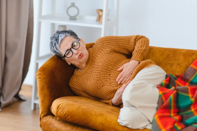 Woman with short gray hair and glasses in an orange sweater lying on a mustard sofa under a plaid blanket, holding her belly with a pained expression