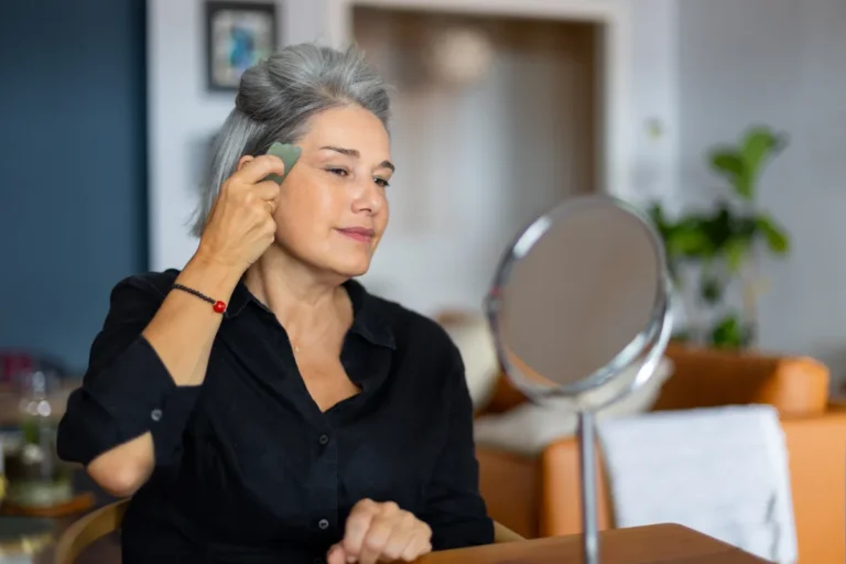 Woman with gray hair in a black shirt sitting at a table and gliding a green gua sha stone along her temple in front of a round mirror