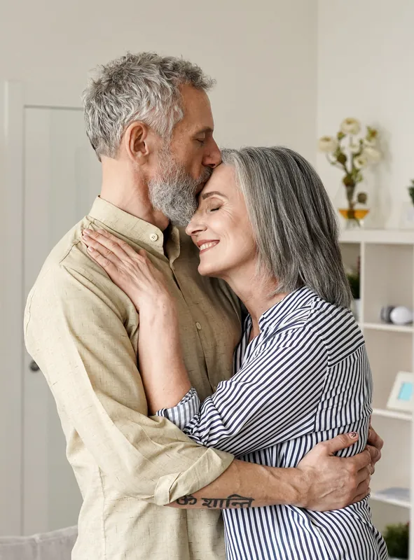 Grey-haired man kissing the forehead of a smiling woman as they embrace at home