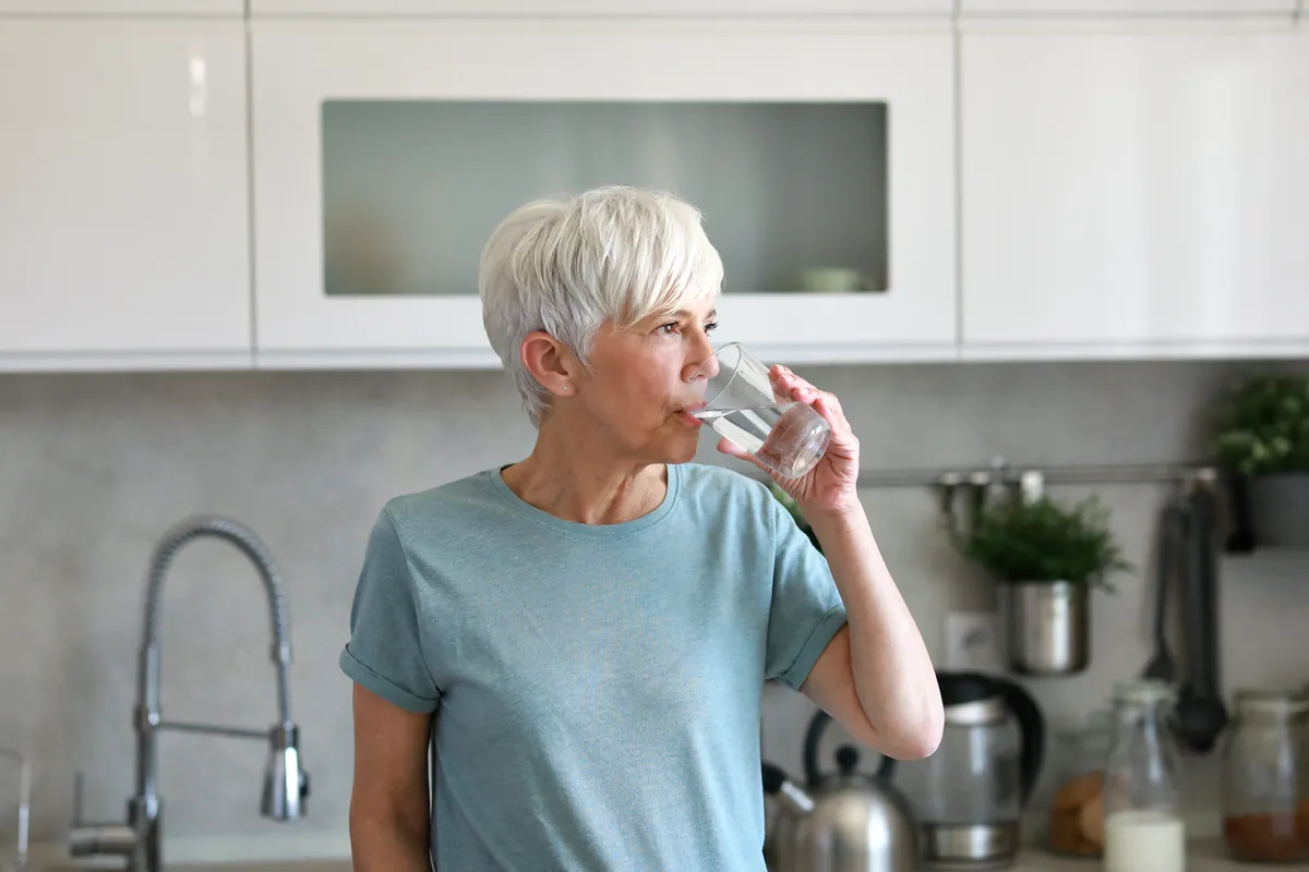 Woman with short grey hair drinking a glass of water in a bright kitchen