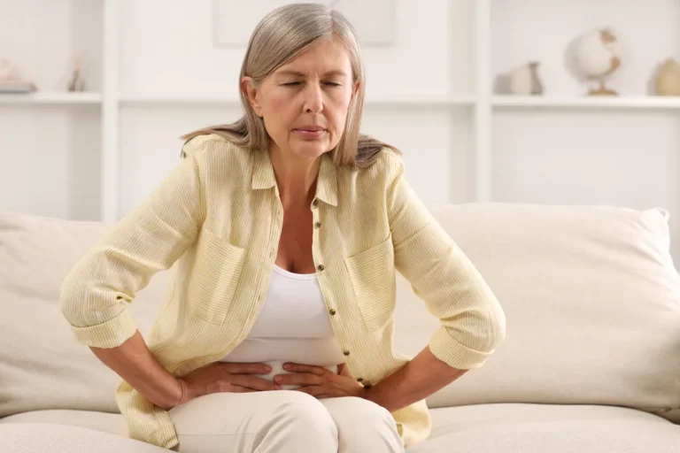 Grey-haired woman in a yellow shirt sitting on a sofa with both hands on her lower abdomen