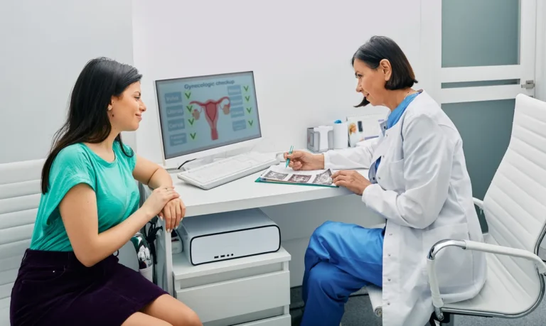 Woman in a teal top talking with a gynecologist at her desk, with a diagram of the uterus on the computer screen