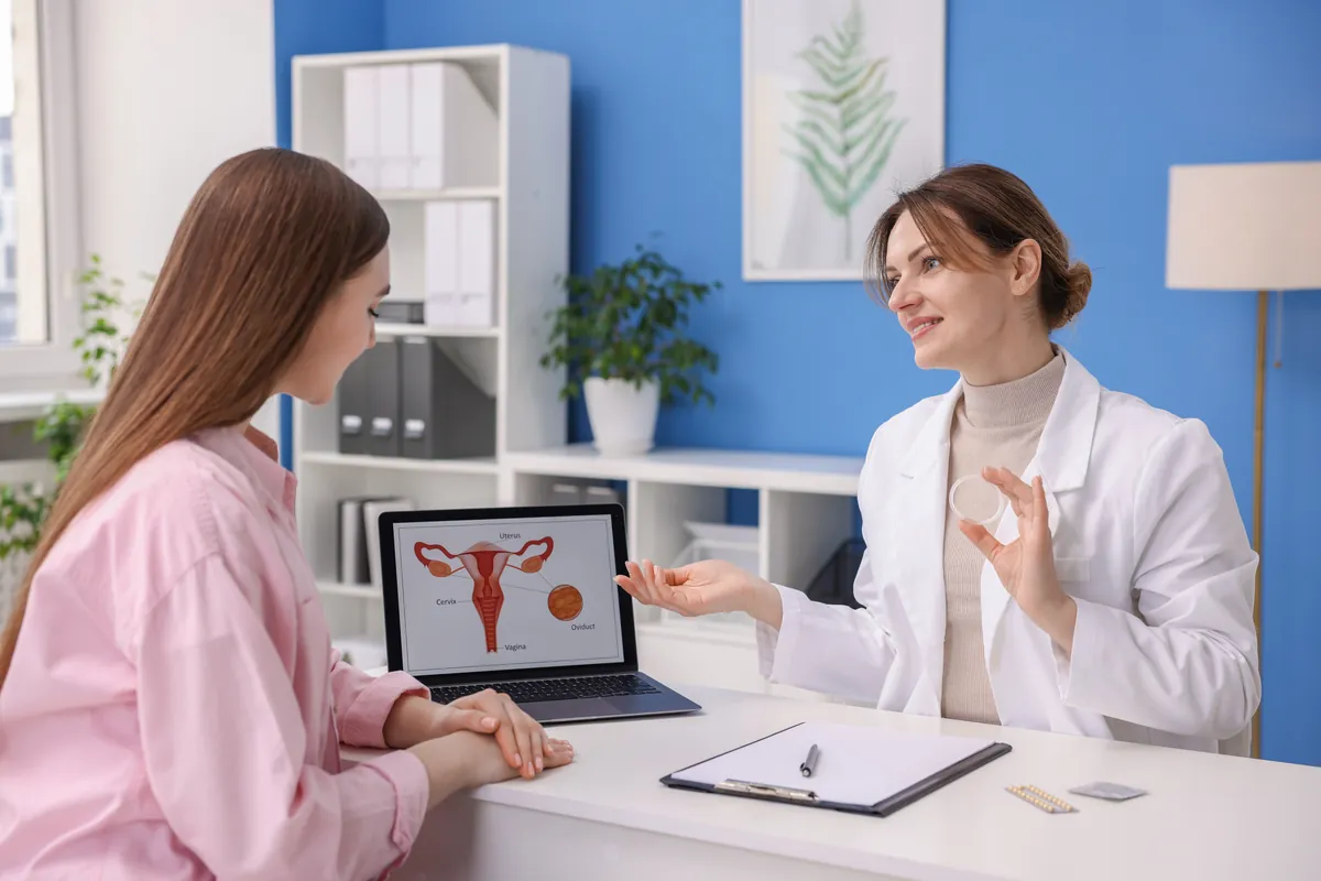 Gynecologist holding a contraceptive ring while talking with a young woman, with a diagram of the uterus on a laptop
