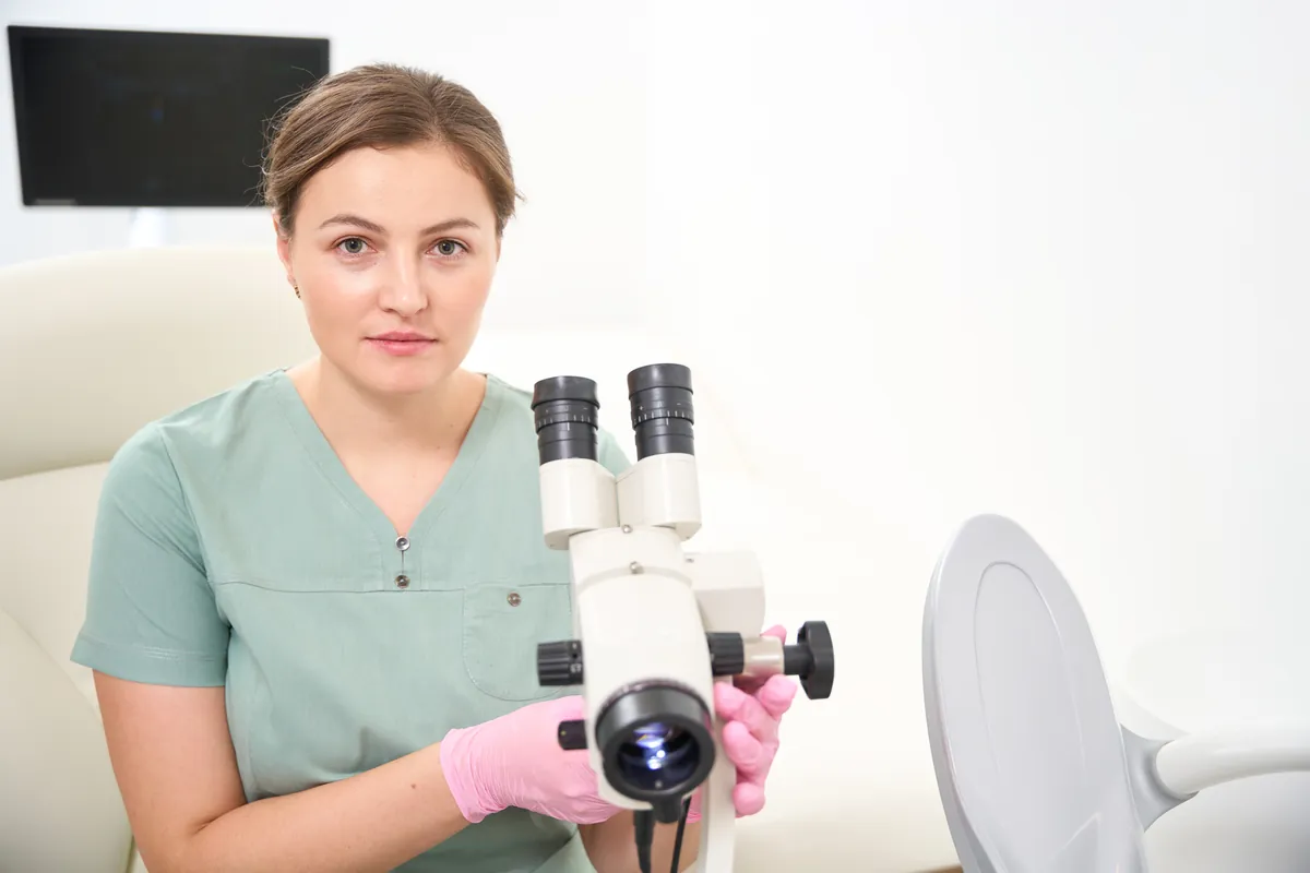 Gynecologist in green scrubs and pink gloves sitting beside a colposcope in an exam room