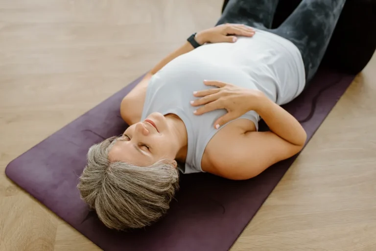 Mature woman with short grey hair lying on a purple mat with her hands on her chest and belly, breathing slowly