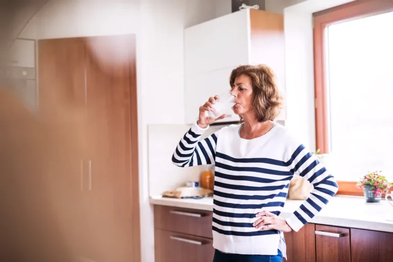 Mature woman in a striped sweater drinking a glass of water in her kitchen, one hand on her hip