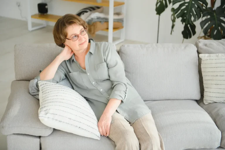 Mature woman with short brown hair and glasses resting on a light gray sofa against a striped cushion, hand behind her neck, looking thoughtfully to the side