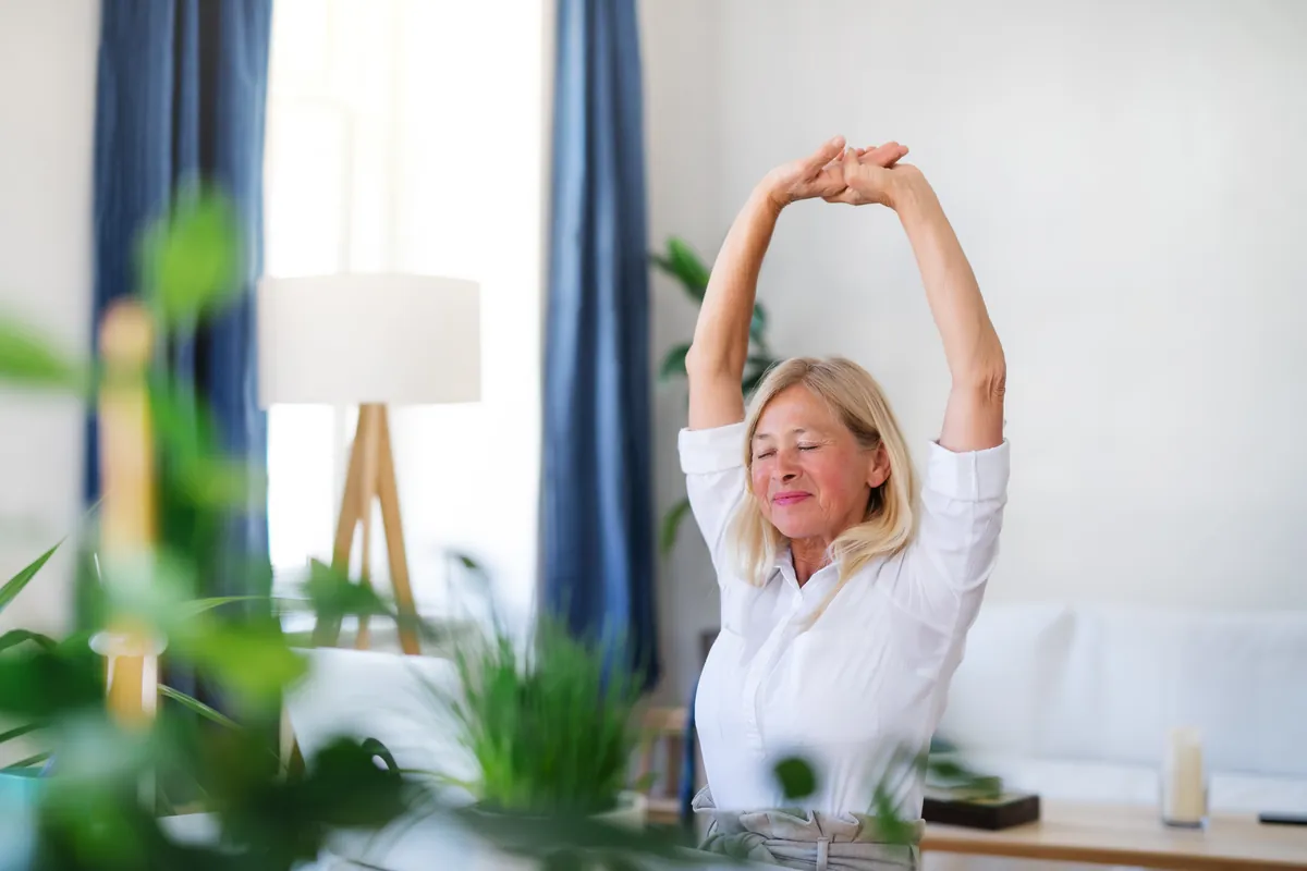 Mature woman with blond hair and a white shirt stretching her arms above her head with her eyes closed, in a bright living room with blue curtains and green plants