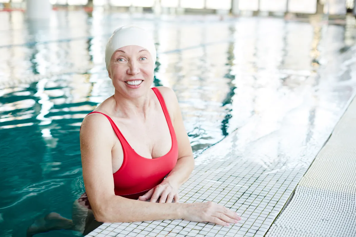 Smiling mature woman in a swim cap resting at the edge of an indoor pool