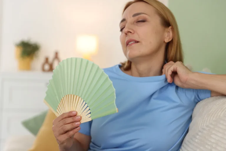 Middle-aged woman in a light blue T-shirt sitting on a sofa at home, eyes half closed, fanning herself with a green paper fan and pulling her collar away from her neck