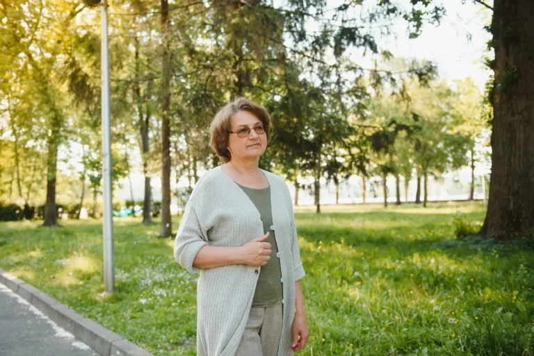 Middle-aged woman with short brown hair and glasses, wearing a light gray cardigan over a green top, standing on a path in a sunny park with tall trees and grass