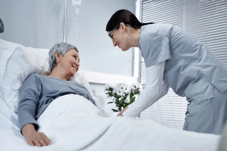 Nurse in light blue scrubs and glasses leaning over a smiling older woman with short grey hair lying in a hospital bed, with an IV bag and white flowers behind them