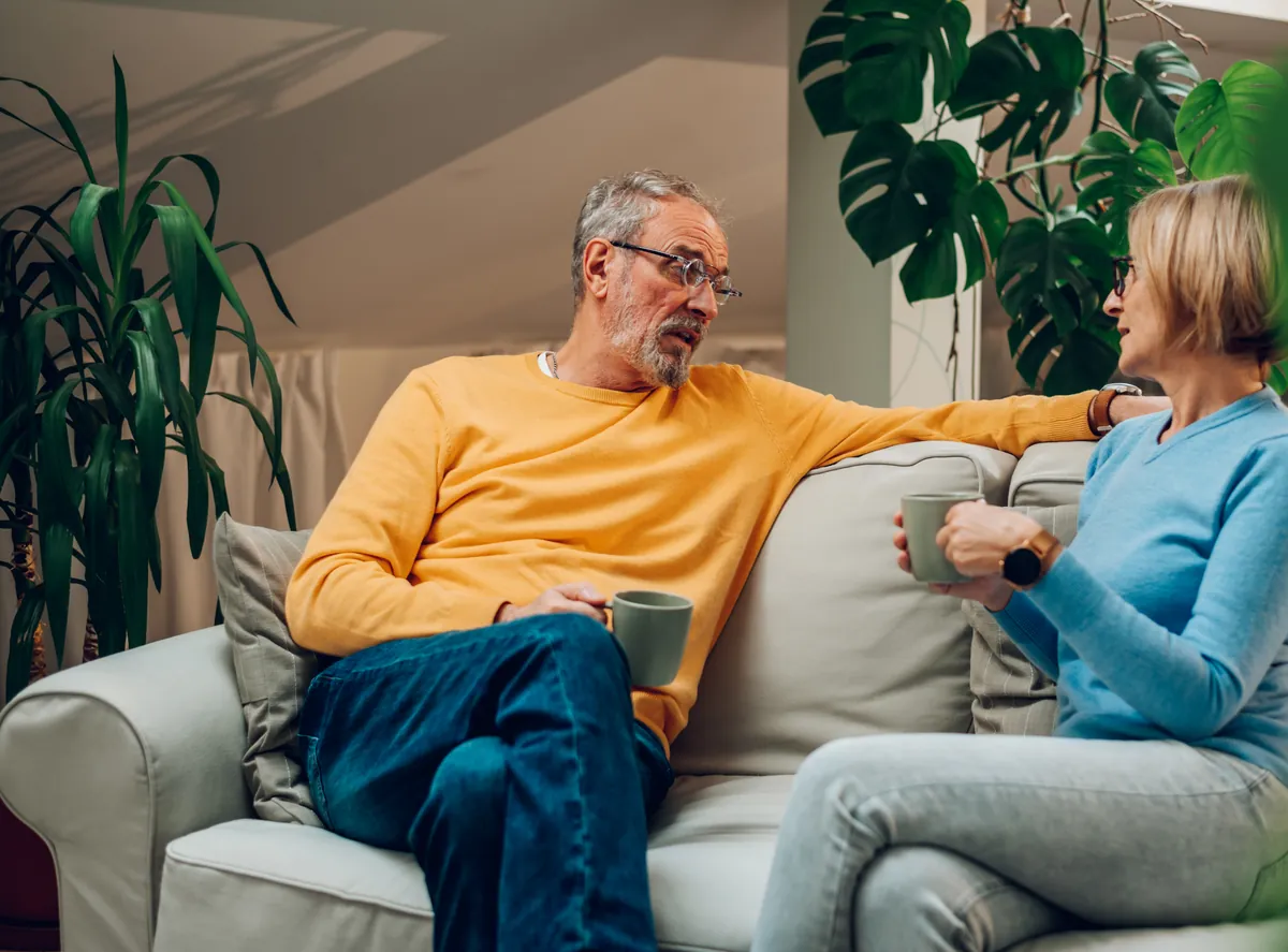 Older couple sitting on a sofa holding mugs and talking to each other