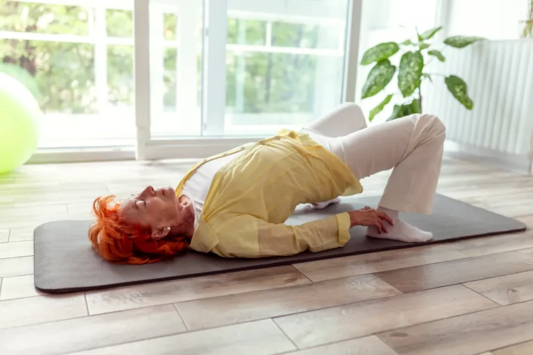 Older woman with red hair, in a yellow top and white trousers, lying on a gray exercise mat on a wooden floor and lifting her hips in a bridge exercise, next to a large window