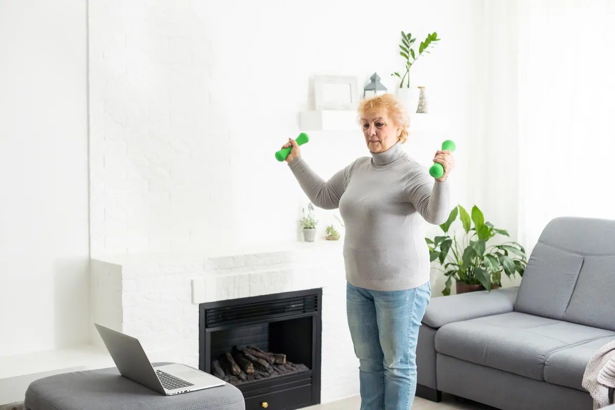 Woman in her sixties in a gray turtleneck sweater and jeans lifting two small green dumbbells to shoulder height in a white living room, with a laptop on a footstool beside her