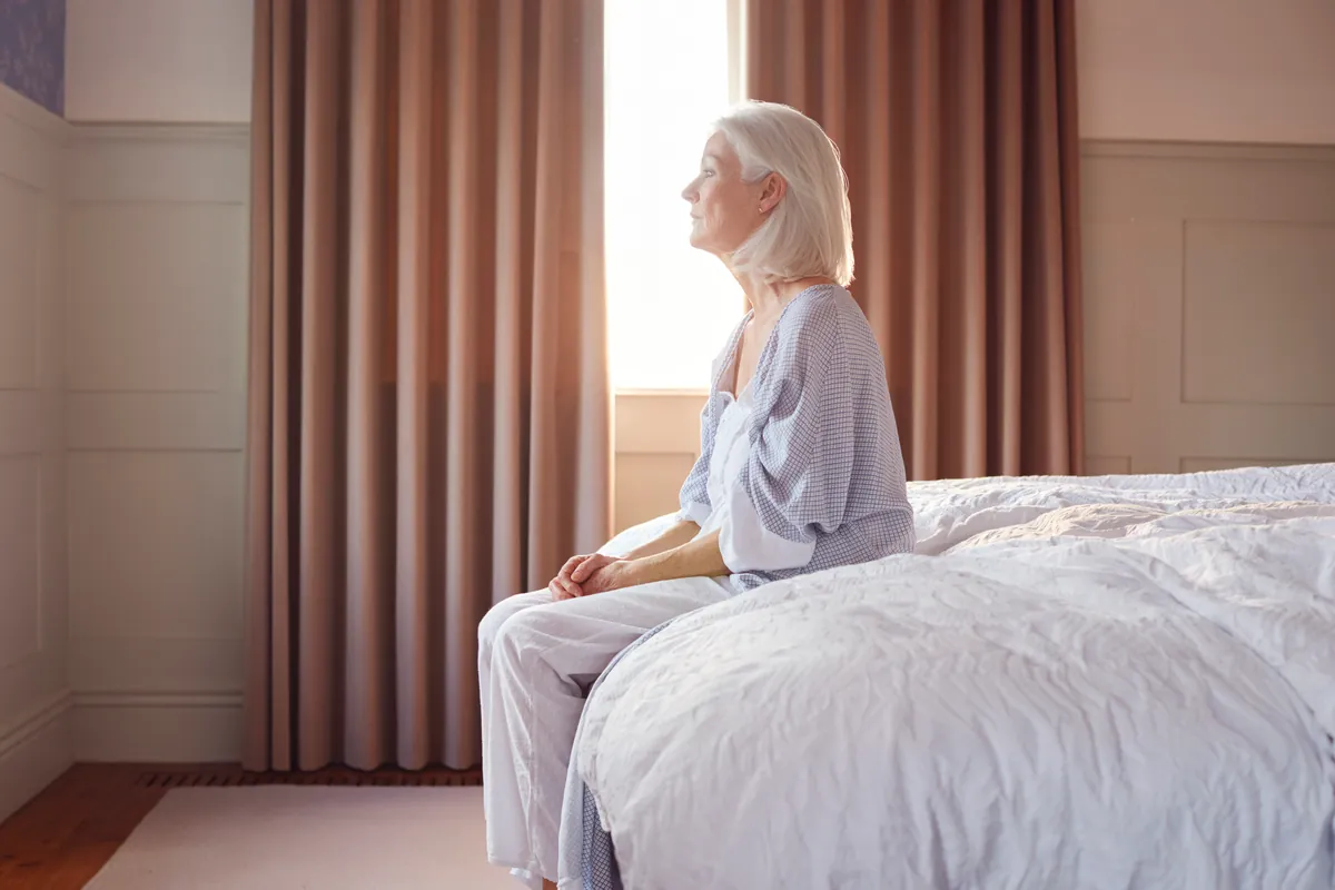 Older woman in pajamas sitting on the edge of her bed before standing up slowly