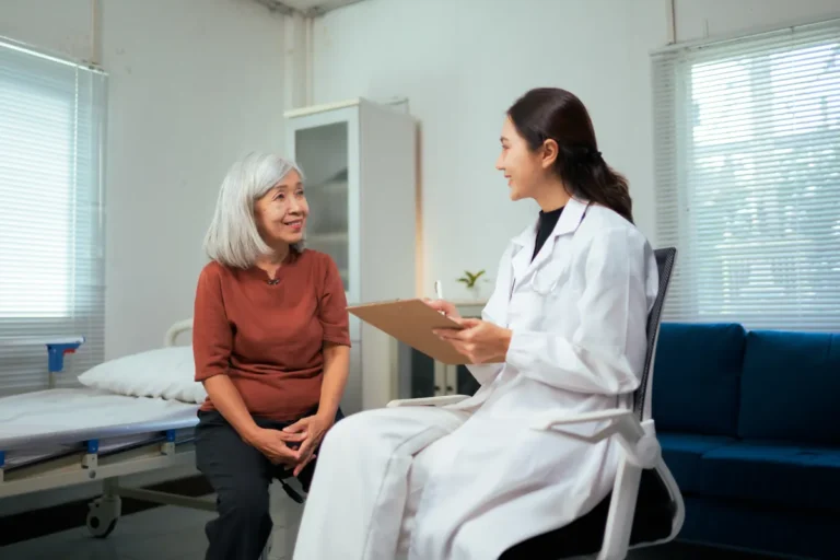 Gray-haired woman in a rust-colored top sitting on the edge of a clinic bed, smiling as she talks with a young female doctor in a white coat who holds a clipboard