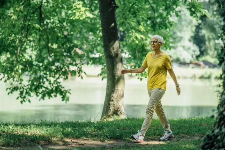 Older woman with short gray hair and glasses, in a yellow T-shirt, beige trousers and white sneakers, walking on a grassy path under trees beside a lake