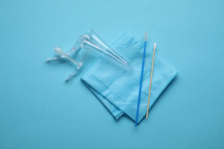 Clear plastic vaginal speculum, blue cervical sampling brush and cotton-tipped swab on a folded blue paper sheet against a blue background