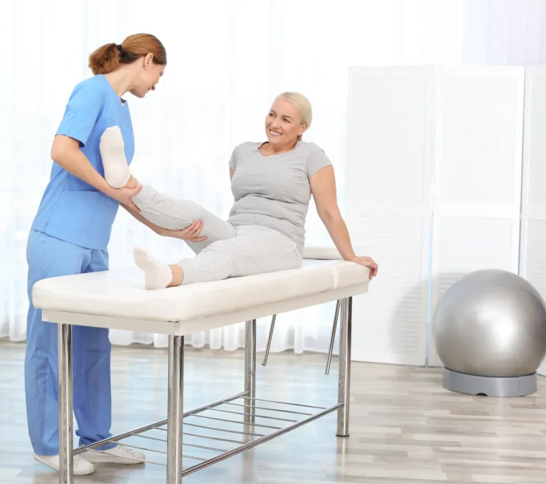 Physiotherapist in blue scrubs guiding a smiling mature woman through a leg exercise on a treatment table