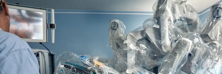 Arms of a robotic surgical system covered in clear sterile drapes in an operating room, with a monitor and a surgeon at the left