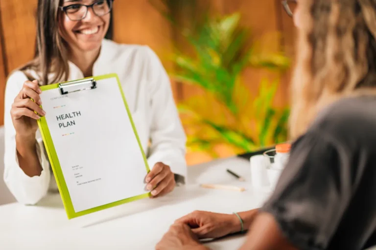 Smiling female doctor in glasses and a white coat holds up a clipboard reading Health Plan to a woman with long hair seated across the desk, seen from behind