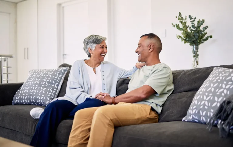 Smiling grey-haired woman and her partner laughing together on a sofa, holding hands