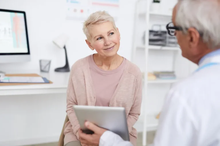 Smiling older woman with short blonde hair and a pink cardigan listening to a grey-haired doctor holding a tablet in a bright clinic office