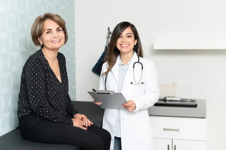 Smiling middle-aged woman sitting on an exam table next to a smiling doctor in a white coat holding a clipboard