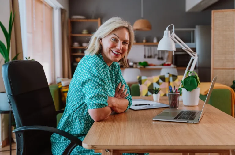 Smiling woman with blond hair in a green patterned dress, arms crossed, sitting at a wooden desk with a laptop and a desk lamp in a home office