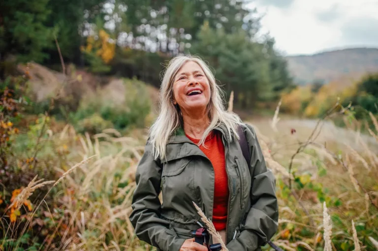 Woman with long gray-blond hair in a green jacket and orange top laughing during a walk through autumn countryside