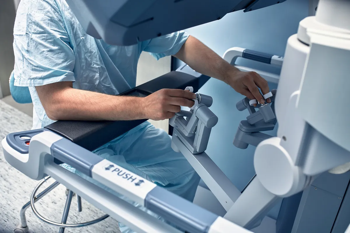 Surgeon in blue scrubs with both hands on the hand controls of a robotic surgery console
