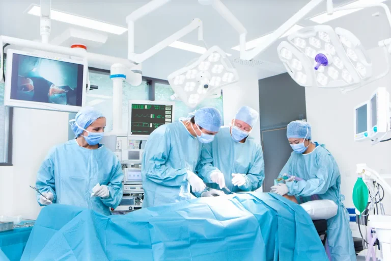 Four members of a surgical team in blue gowns, caps and masks work over a draped patient in a bright operating room with overhead lights and wall monitors