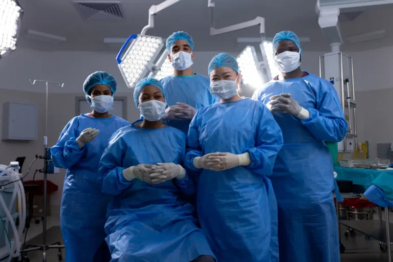 Five members of a surgical team in blue gowns, caps, masks and gloves stand together under the lights of a hospital operating room