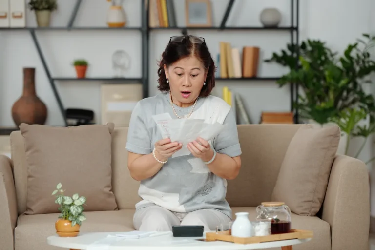 Surprised older woman sitting on a beige sofa reading a handful of bills