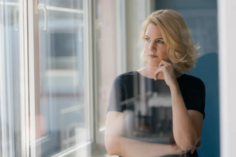 Blonde woman in a dark navy top standing by a window, resting her chin on her hand and looking outside thoughtfully